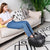 A young woman relaxes on a beige sofa, using her smartphone with her feet in a black Renpho Shiatsu Foot Massager Pro +. The room has a modern look with decorative cushions and a patterned rug.