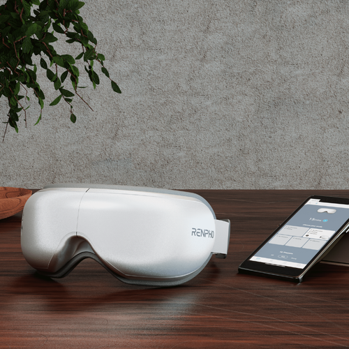 A personalized Renpho Eyeris Smart Eye Massager is sitting on a wooden table next to an eyeglasses.