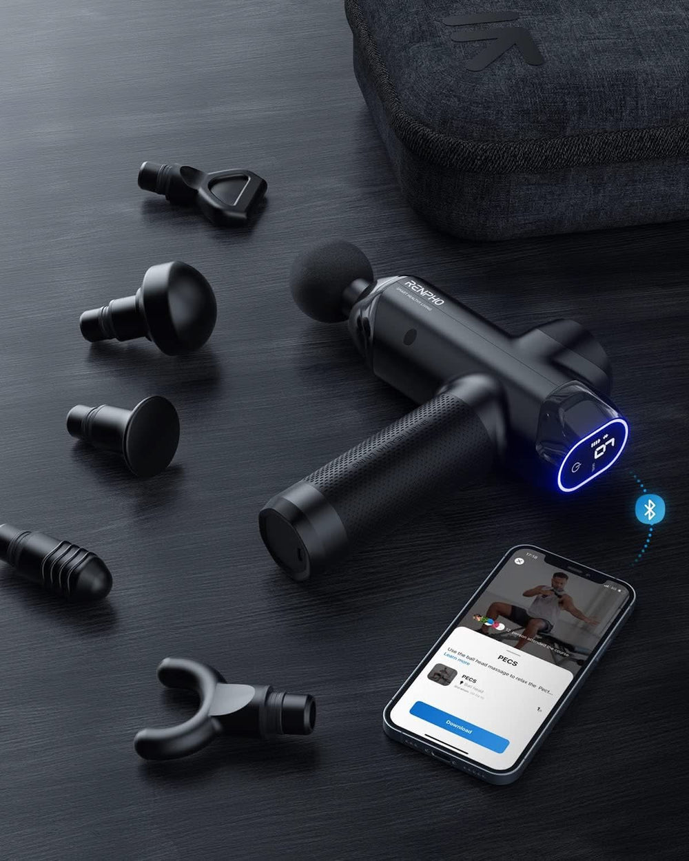 A black Power Massage Gun with a textured handle lies on a dark table, surrounded by various detachable massage heads. Nearby, a smartphone displays an app with a workout video and a Bluetooth icon. A soft carrying case is in the background. The Renpho Power Massage Gun features noise reduction technology ideal for muscle soreness recovery.