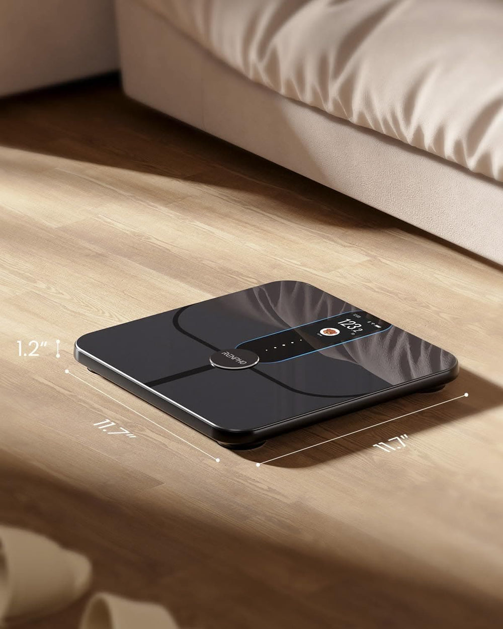 A modern digital Renpho Elis Nova Smart Scale with a sleek black design, located on a wooden floor next to a bed. The display on the scale shows "1.2" and has illuminated indicators for