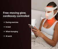 Woman in bed using her phone while wearing a cordless LED face mask, for use in bed, at work or during exercise