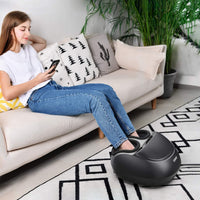 A young woman relaxes on a beige sofa, using her smartphone with her feet in a black Renpho Shiatsu Foot Massager Pro +. The room has a modern look with decorative cushions and a patterned rug.